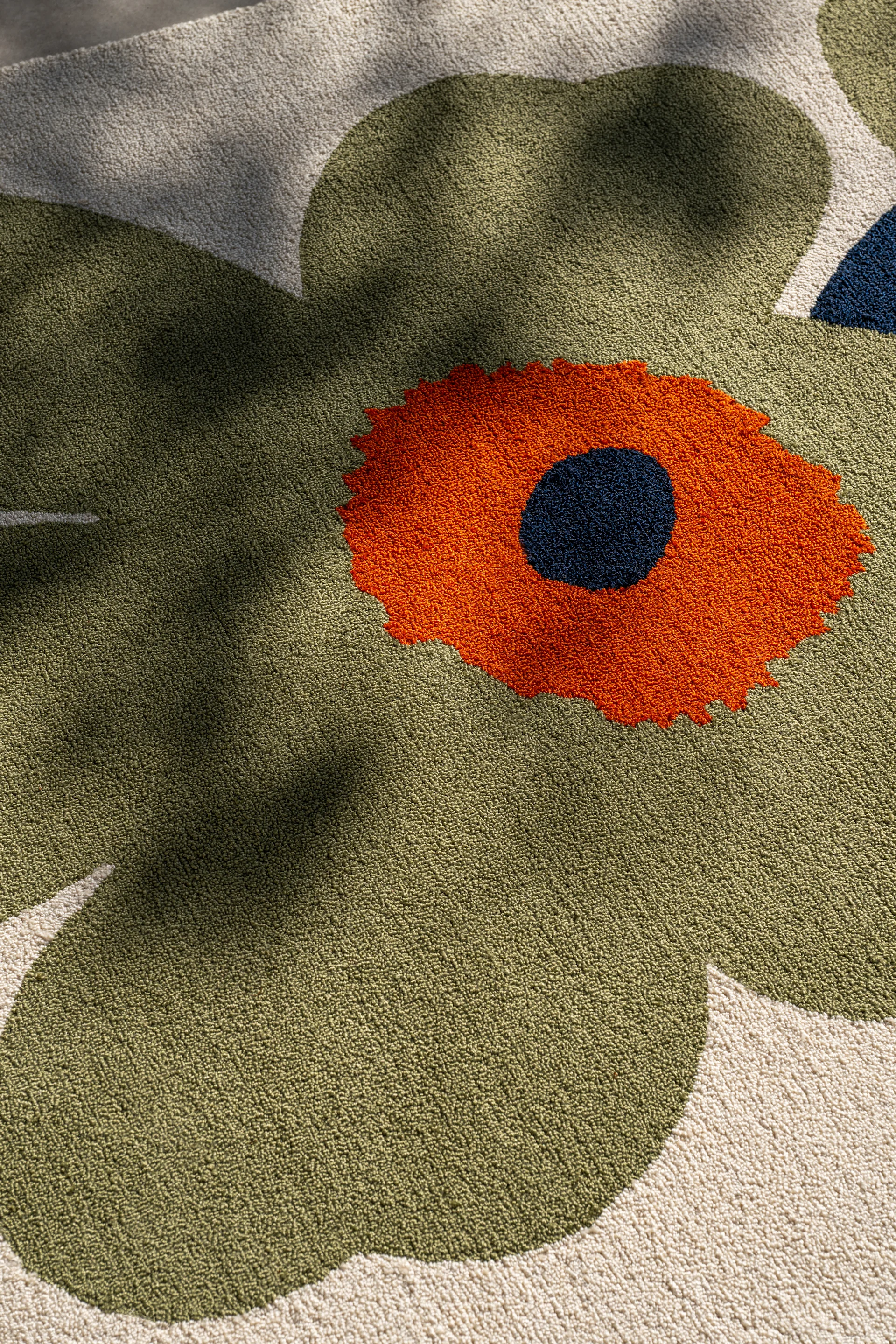 Gros plan d'un tapis avec un grand motif floral stylisé. Pétales vert olive, centre orange et point bleu foncé sur fond crème.