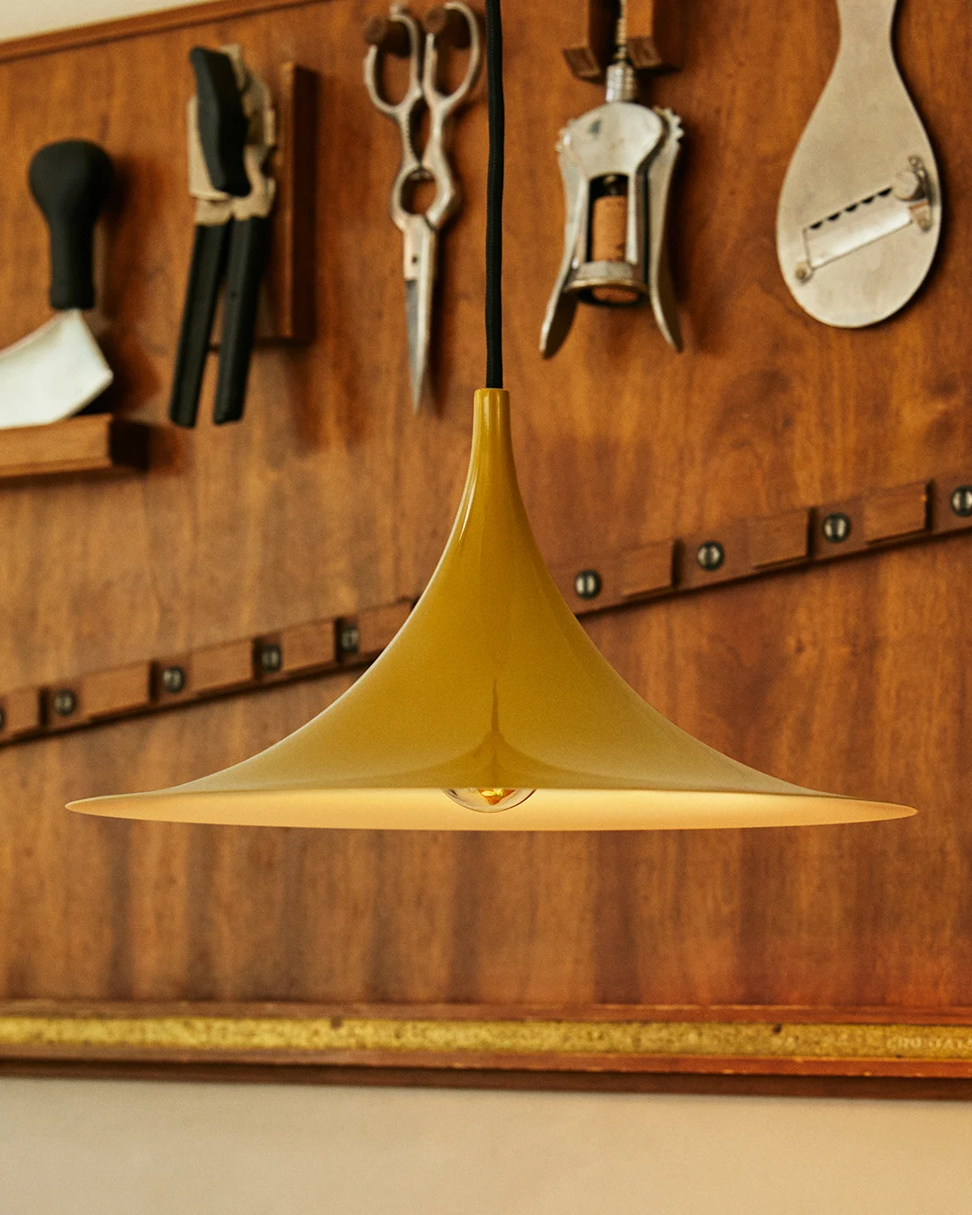 Lampe suspendue dorée en forme de cloche avec ampoule visible, devant un mur en bois avec outils.