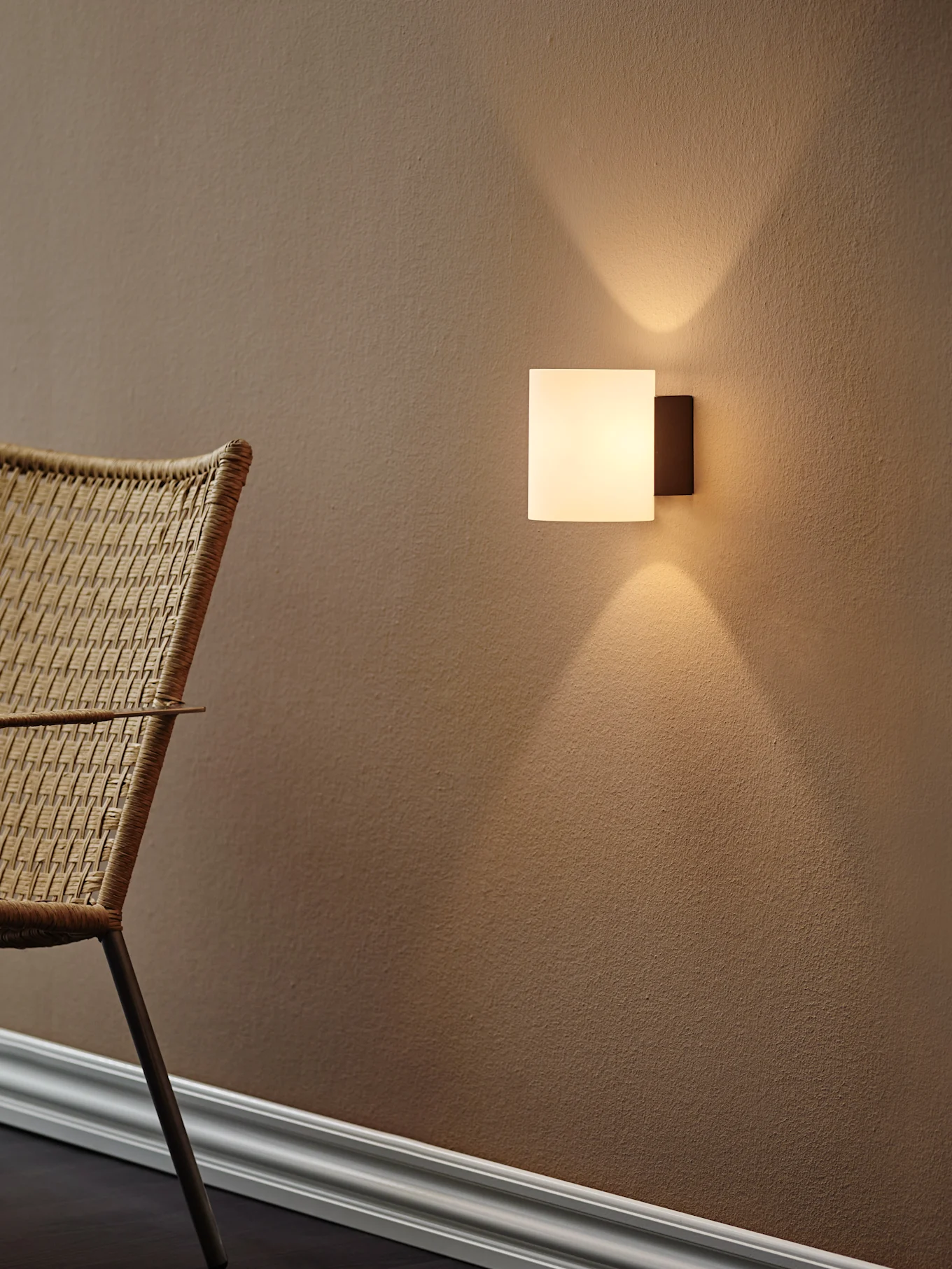 Applique murale carrée moderne projetant une lumière chaude vers le haut et le bas sur un mur texturé brun, avec une chaise tressée à gauche.