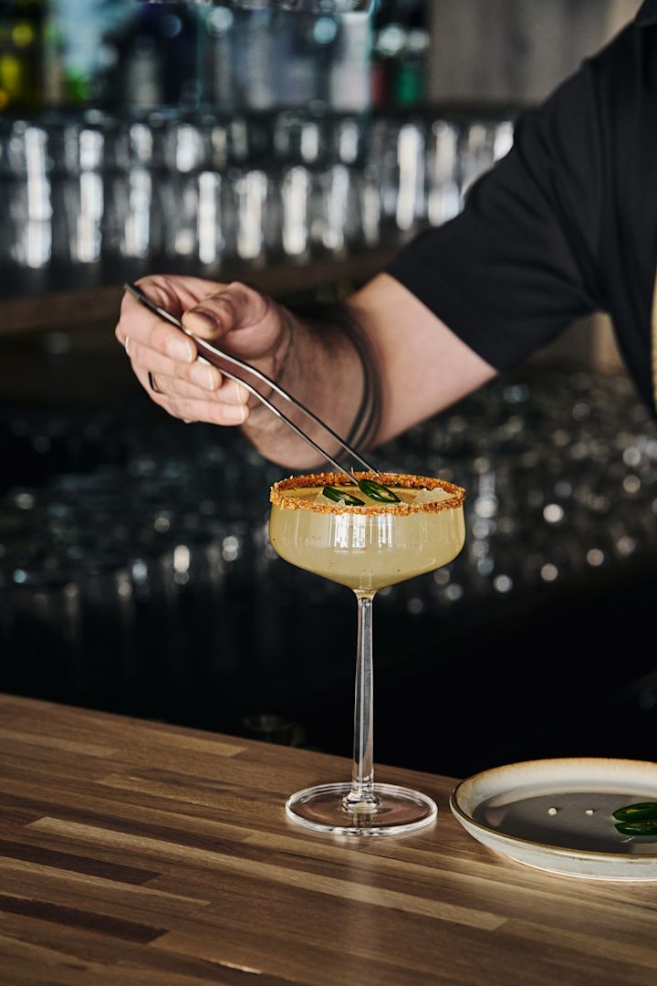 Parfait pour les cocktails sans alcool : le verre à cocktail Essence d'Iittala avec boisson, bord décoré et garni de tranches de jalapeño à l'aide d'une pince.