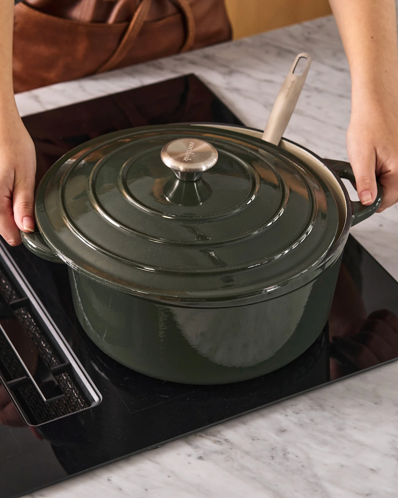 Personne plaçant une cocotte en fonte vert foncé avec un bouton métallique sur une plaque à induction noire.