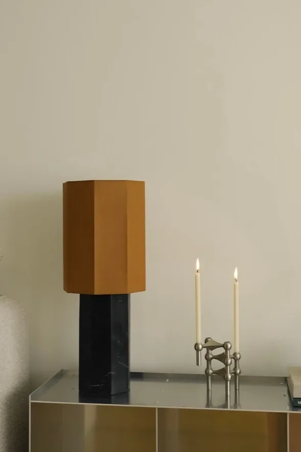 Une lampe de table moderne avec un abat-jour hexagonal marron et un socle en marbre noir, à côté d'un bougeoir modulaire argenté surmonté de deux bougies blanches allumées.