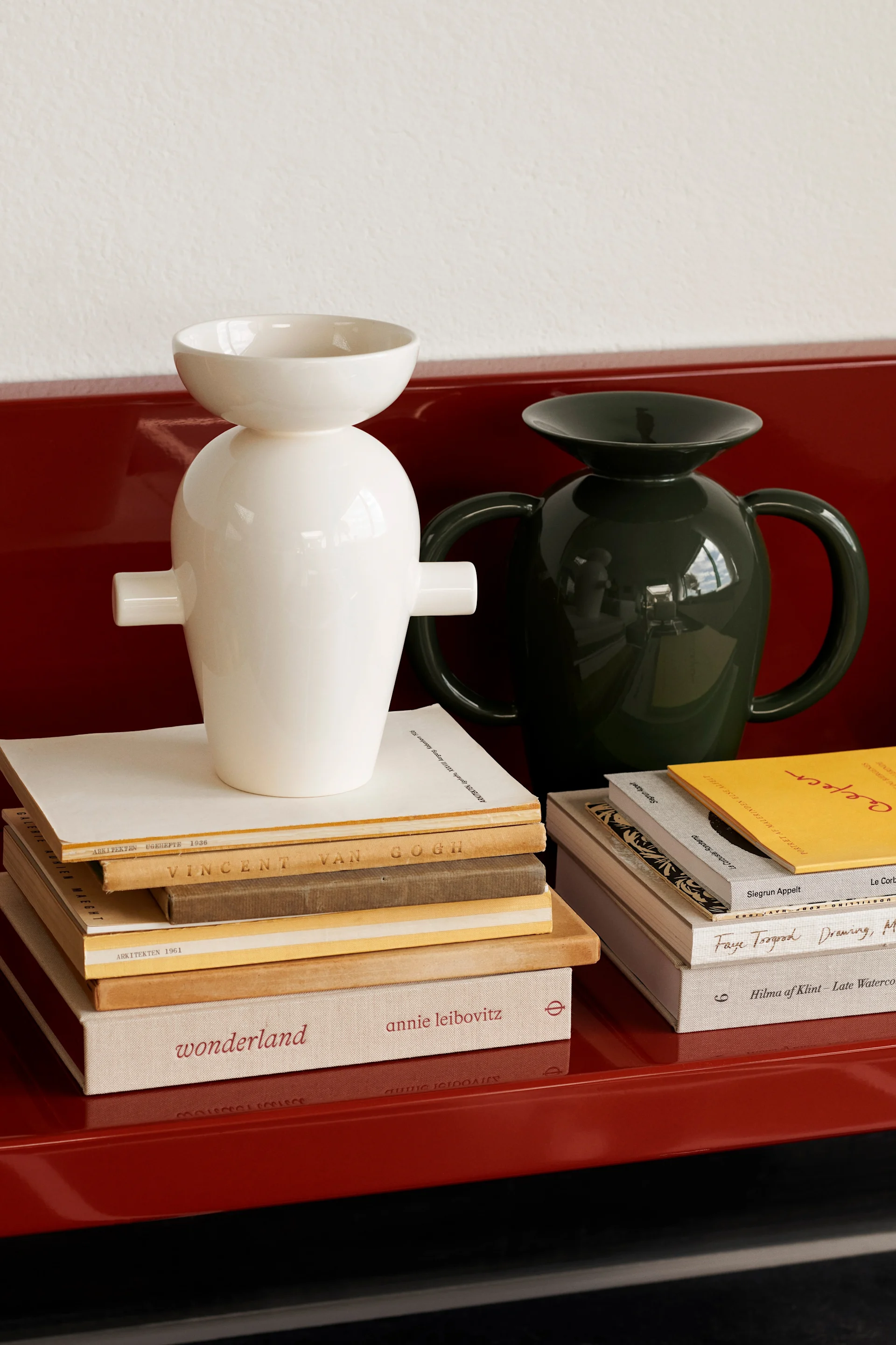 Deux vases en céramique brillants, un blanc et un vert foncé, posés sur des piles de livres sur une surface rouge.