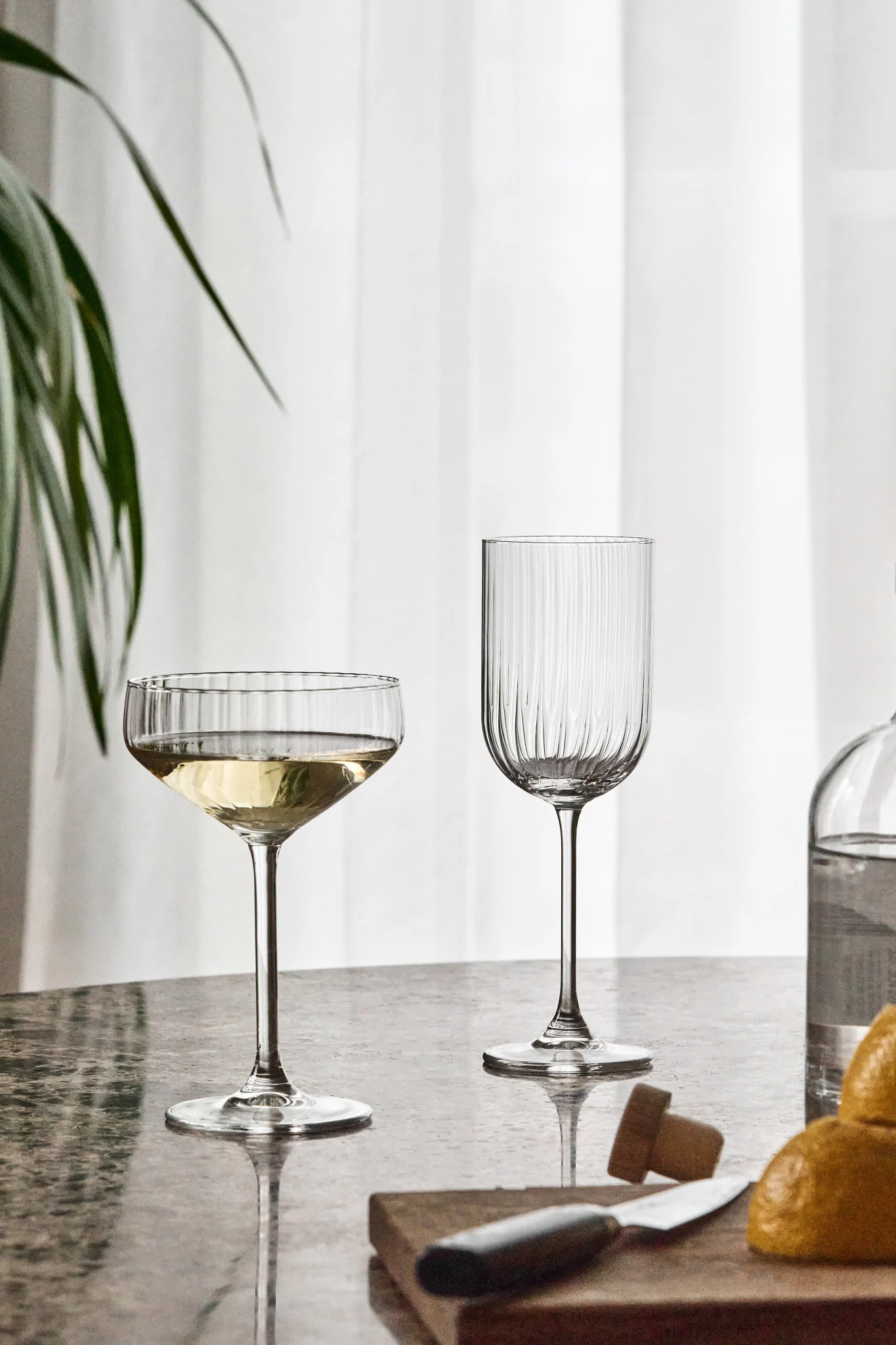 Deux verres cannelés sur une table réfléchissante, l'un rempli de liquide blanc. Citrons, couteau et bouteille flous au premier plan.