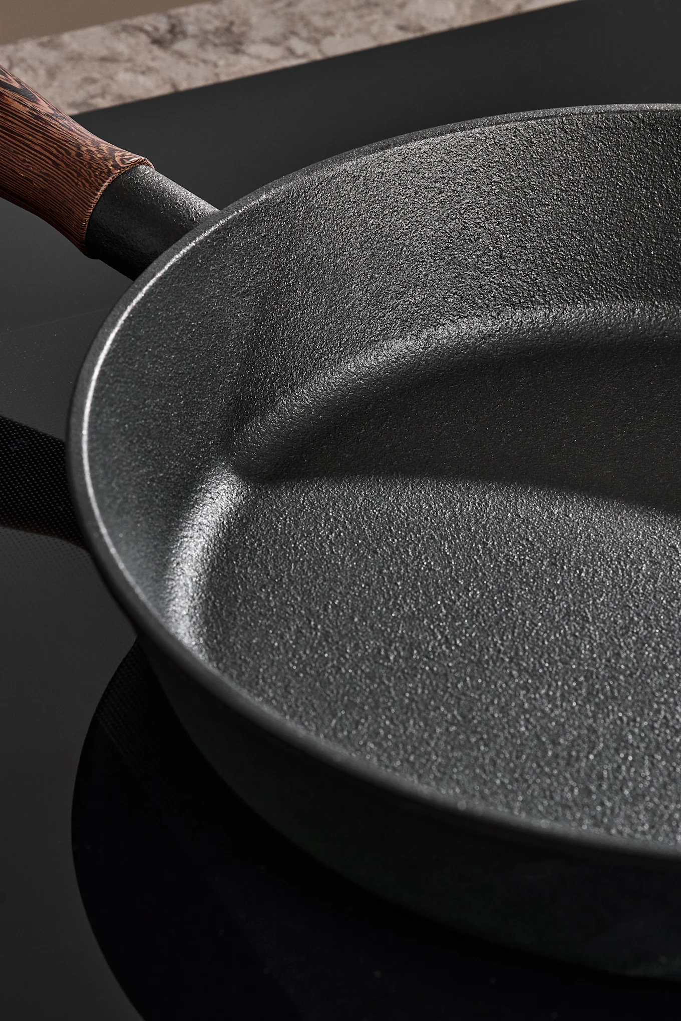 Gros plan d'une poêle sombre et texturée avec un manche en bois sur une plaque de cuisson noire.