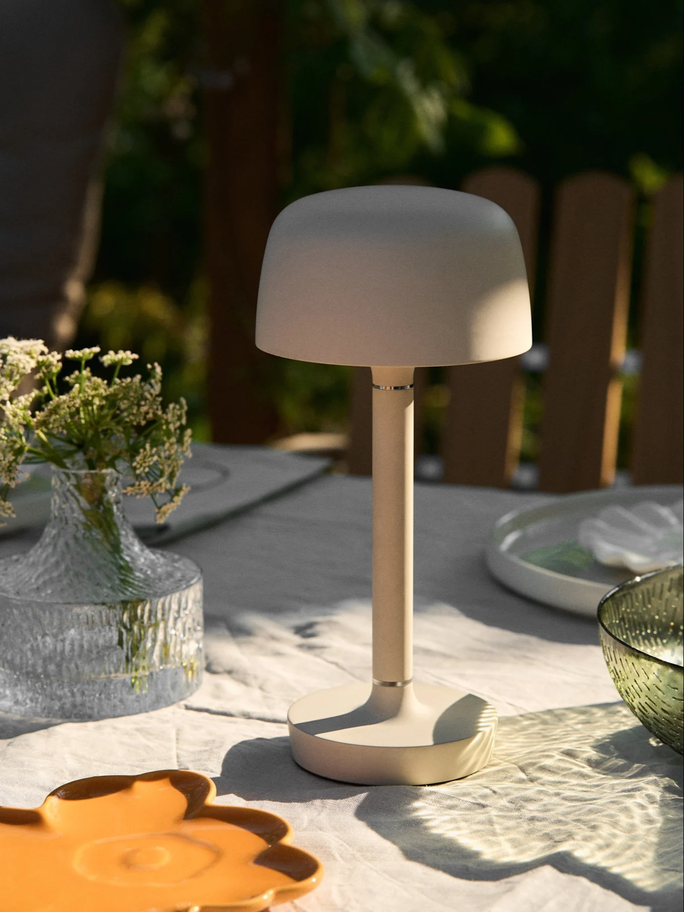 Lampe de table portable beige sur une table d'extérieur avec un vase de fleurs et un soleil tacheté.