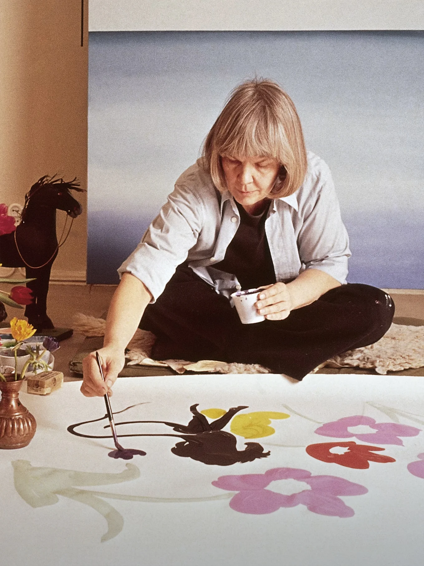 L'artiste Marimekko Maija Isola assise par terre peint de grandes fleurs colorées sur un tissu blanc.