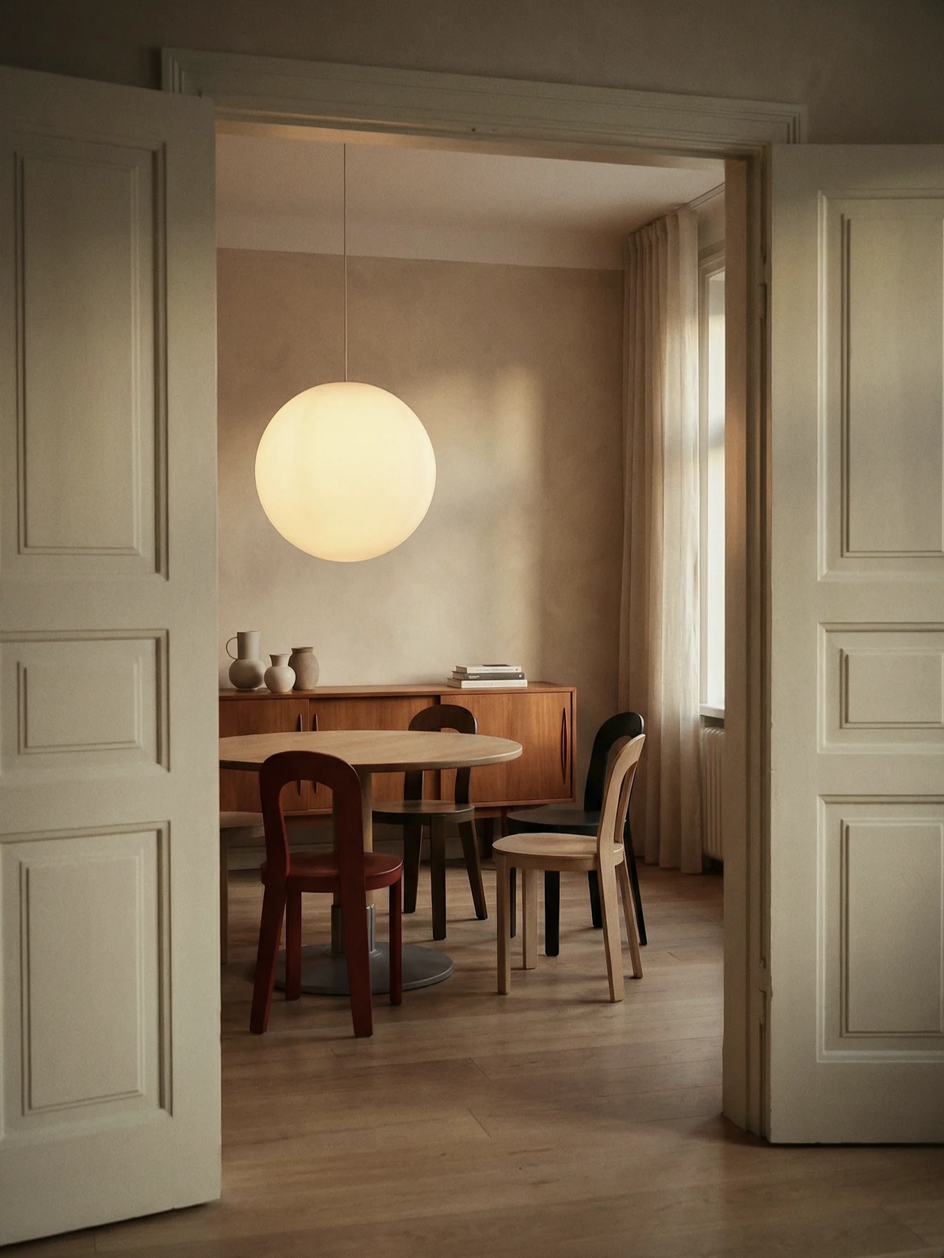 Salle à manger par porte ouverte, avec table ronde en bois, chaises, grande suspension sphérique et buffet.