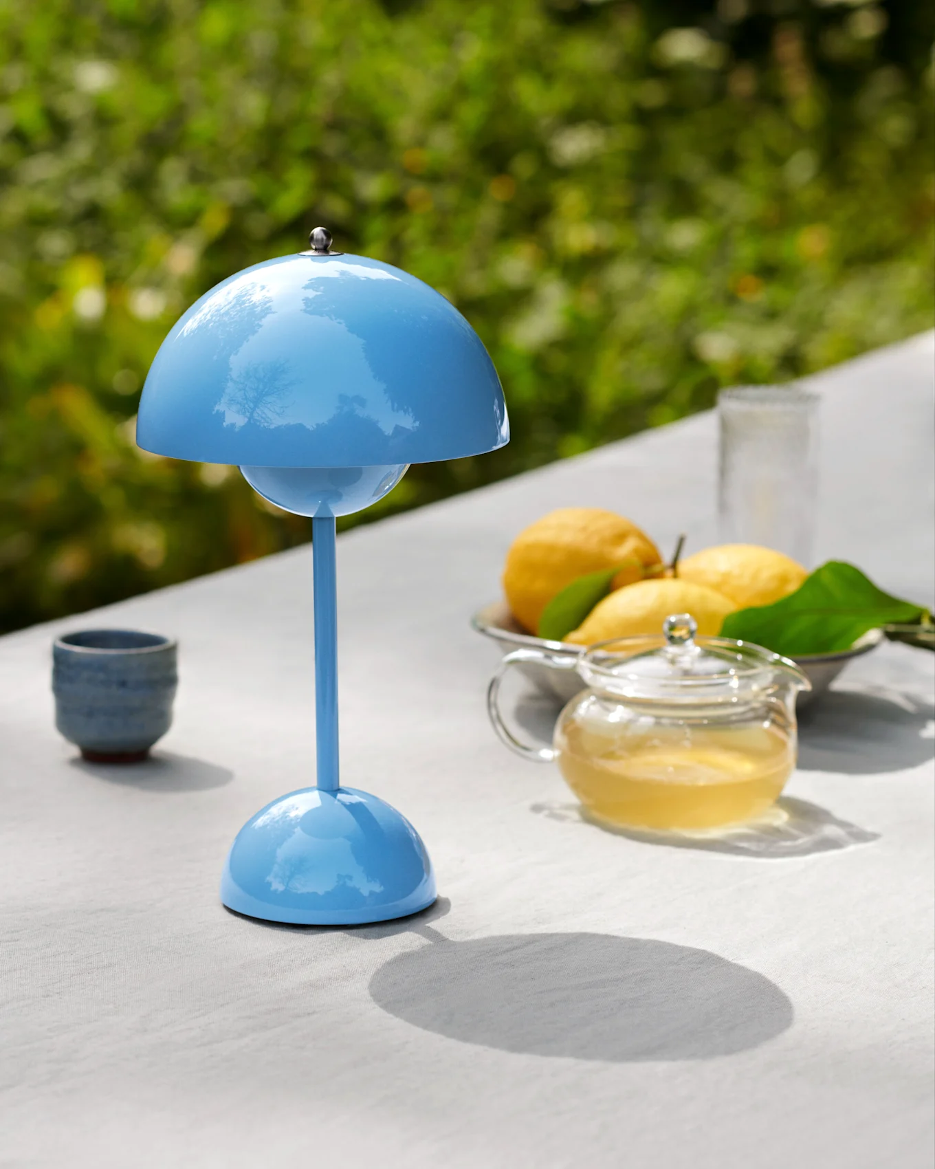 Une lampe de table bleue en forme de champignon sur une table extérieure avec une théière, des citrons et une tasse.