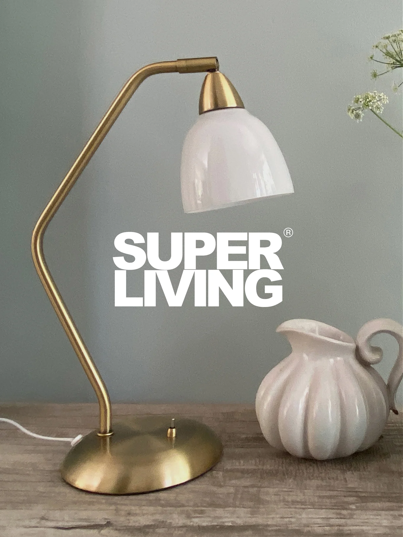 Marque SUPER LIVING sur une lampe de bureau en laiton avec un abat-jour blanc et un pichet cannelé blanc sur une table en bois.