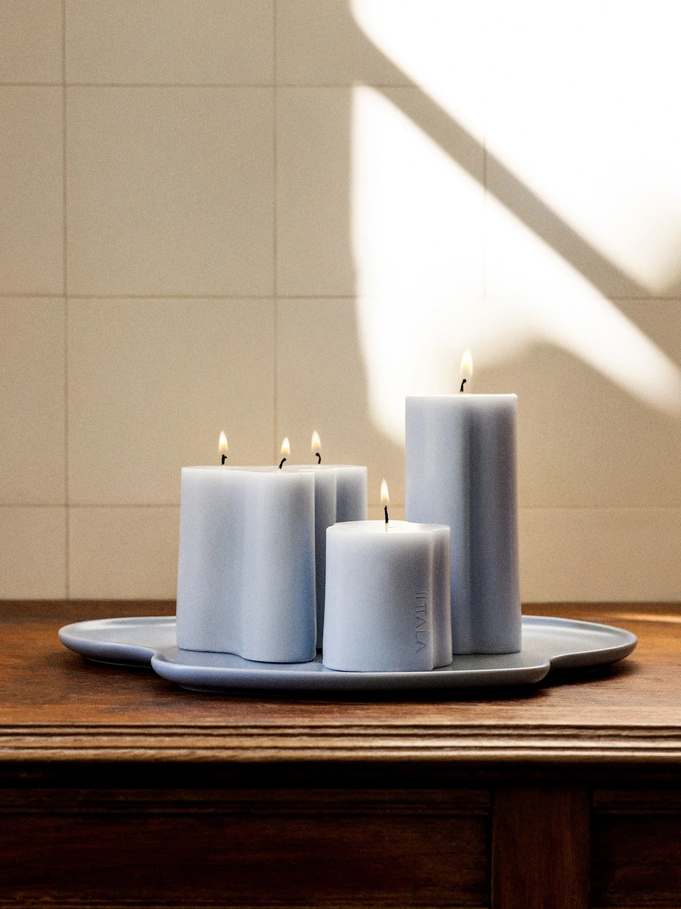 Quatre bougies allumées bleu-gris clair de différentes formes et hauteurs sur un plateau assorti de forme irrégulière sur une surface en bois foncé.
