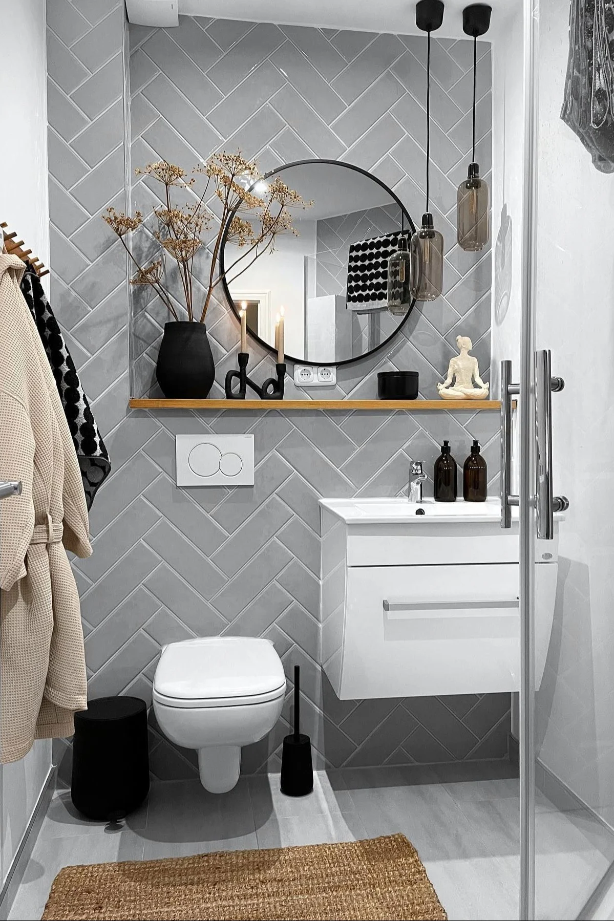 Salle de bain moderne avec carrelage mural à chevrons gris clair, toilettes blanches, meuble-lavabo, miroir rond et étagère en bois décorée.