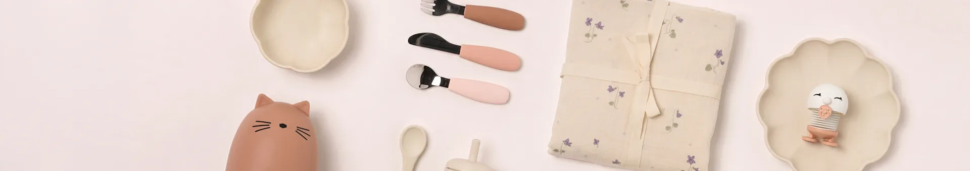 Vaisselle bébé à plat: assiettes beiges, biberon chat, couverts roses, couverture florale, jouet œuf.